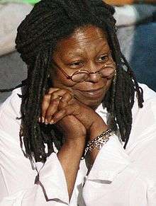 Middle-aged African-American woman with dreadlocks, looking away from the camera and smiling with her right cheek rested upon her folded hands.