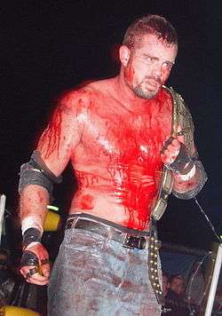 Adult white male covered in blood wearing blue jeans and wrestling gear while holding a gold championship wrestling belt.