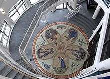 An aerial view of a mosaic depicting five people working, mining, farming -- all radiating out from the center like a flower. The are also tools of various trades like a hammer and a scythe surrounding the flower-like part.
