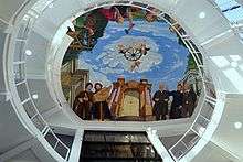 A view looking upward at a ceiling mural. It shows men of different eras hammering, reading a map, pointing in the distance. The view is framed by railings on all sides.