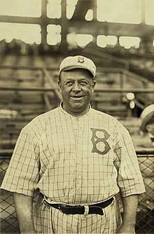 A man, wearing a baseball cap with a "B" in the center and a white baseball uniform with a square-pattern design and the Brooklyn Dodgers stylized "B" logo on the left breast, looks forward smiling.
