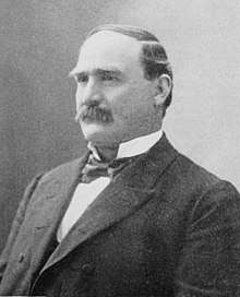 A man with receding, black hair and a thick, black mustache wearing a black jacket and tie and white shirt