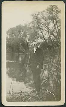 Cartheuser smiling at camera while standing at the edge of a body of water dressed in a suit and holding a hat.