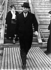 A middle-aged man in a dark coat and top-hat strides confidently along a boardwalk at the base of a set of steps. A well-dressed woman is on the steps behind him, while a third person, a man, approaches the steps.
