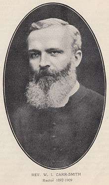 Oval portrait photo of Carr Smith, a strong-faced man with large square forehead, deep-set dark eyes, and a bristly beard partly grey and partly white.