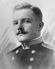 Head and shoulders of a white man with a neatly trimmed mustache wearing a military jacket with shoulder boards and two columns of buttons down the chest.