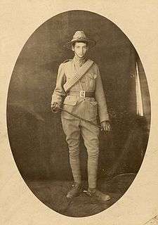 Full-length studio portrait of a young man in khaki military uniform. He has a broad brim hat on, and is holding a riding crop.
