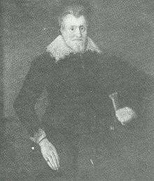 Formal portrait of Sir William Davenport, standing. He has a long pale face, hair combed back from his forehead and a trim full-face beard. He wears dark clothing with a falling collar and cuffs bordered with needle lace and a ring on his finger