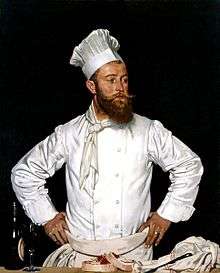 A chef with a bushy brown beard and waxed moustache wearing his white kitchen attire and a tall chef's hat stands with his hands on his hips as if he has been interrupted in his work.