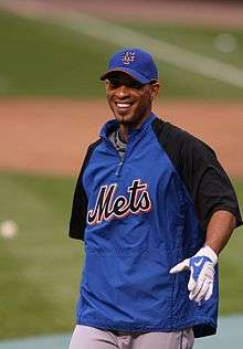 A smiling brown-skinned man wearing a blue warmup jacket with "Mets" across the chest and a blue baseball cap