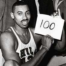 Wilt Chamberlain, an African American man, is shown sitting down in his Philadelphia Warriors jersey while holding up a piece of paper with the number 100 written on it. The photograph was taken directly after the game and is in black and white.