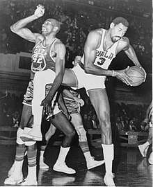 A basketball player, wearing a jersey with the word "PHILA" and the number 13, is holding a basketball while another basketball player, wearing a jersey with the word "RIP CITY" and the number 42, is standing next to him.