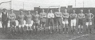 A black-and-white photograph of a row of twelve men standing in front a football goal. The man furthest to the left is dressed in old-fashioned, but smart work clothes; the remainder are wearing football attire. While the shorts and socks worn by these men vary, the dark shirts bearing a "W" are the same except for one, worn by the man standing in the middle of the photograph, which is white.