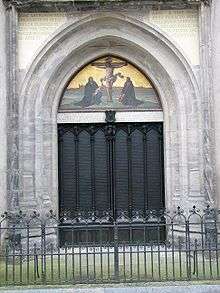 Two large black church doors with a crucifixion scene painted above with Luther and Melanchthon kneeling