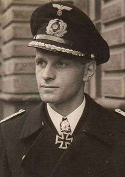 The head and shoulders of a young man, shown in semi-profile. He wears a peaked cap and black naval coat, a white shirt with an Iron Cross displayed at the front of his shirt collar.