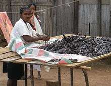 A Malagasy woman sorts vanilla pods heaped on a wooden table outdoors