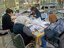Four women, one in a wheelchair and two wearing headscarves, sitting around a table assembling mosaics with tools and materials