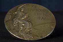 A tarnished gold medal featuring a person in a toga, and the words "XI Olympiade Berlin 1936".