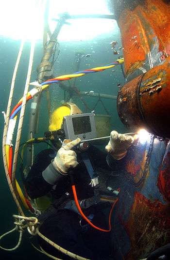 Helmeted surface-supplied diver using a coated electrode to arc-weld a steel patch to the underwater hull of a landing craft.