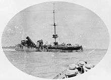 A badly damaged ship sits in the water, while in the foreground sailors in another vessel watch on.