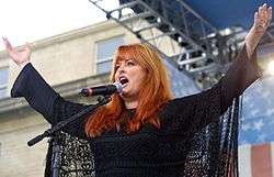 A woman with long red hair wearing black, singing into a microphone while raising both her arms