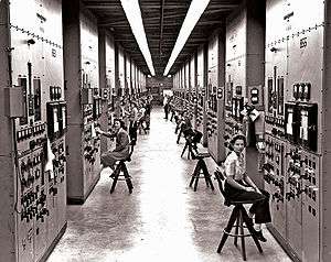 A long hallway, with bulky computers reaching from the floor to the ceiling, covered in switches and dials. Women sit on chairs operating the machinery.
