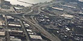 A series of highway ramps with multiple cars on them. A body of water is next to them and they are surrounded by buildings