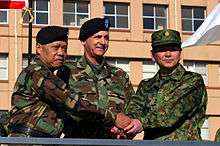 Two American generals wearing battle dress uniforms, and one Japanese general wearing a camouflage field uniform, all shaking hands.