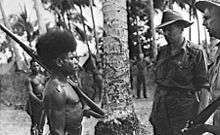 A shirtless Papuan man stands at attention with rifle at slope. Two Australian soldiers wearing shirts and slouch hats stand facing him.
