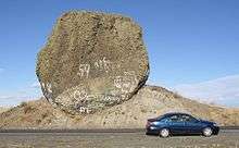 This photo shows an automobile passing in front of a rock which is essentially fully exposed. The rock has a rough, dark surface indicating it is weathered basalt and is roughly circular in exposed cross-section. The rock is immediately adjacent to a roadway – the road cut removed much of the earth from one side of it exposing it &ndash; from the excavation it is evident that the rock sits on a mound of glacial till. The rock is approximately 2 times the length of the car (i.e., ≈9 meters) in one direction and 5 times the height of the car in the other direction (i.e., ≈9 meters). Since the rock has not tipped onto the road and no structural support is provided, it must be approximately as deep as it is wide and high. Since the density of basalt is 3 grams per cubic centimeter, this puts the mass of the rock at about 400 to 500 metric tons (consistent with the references).