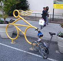 A bike rack shaped like a stylized bicycle with a rider with hair streaming in the wind. It is painted yellow and set in tarmac at the edge of a road between two large concrete plant pots. A folded bicycle is parked there and an abandoned chain hangs from it. Behind, two women walk past on the sidewalk in front of black iron railings.
