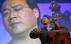 A man wearing glasses and playing the cello with his eyes closed with his image projected in the back.