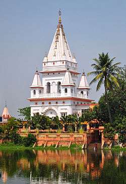 A white ornate structure with a pyramidal pointed dome standing on the bank of a pond and surrounded by trees