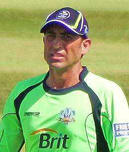 A man in parrot-green shirt and blue cap standing in a cricket ground