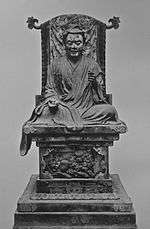 Frontal view of a cross-legged seated statue placed on a throne. His right arm rests on his leg, the left arm is bend and slightly raised.