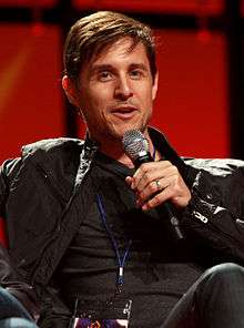 A middle-aged, Caucasian man with short, dark hair, wearing a black jacket and speaking into a microphone