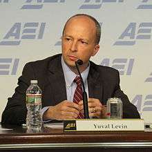 A man with close-cropped, receding hair, wearing a suite, looking intently slightly to his right. He is sitting at a table with a microphone against a blue, repeating ARI logo.