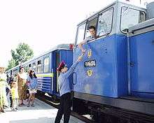 Blue train at a station, with the engineer passing a stick to employee on platform