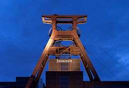 A orange metal tower with several flywheels above a building with Zollverein written in golden gothic script letters.