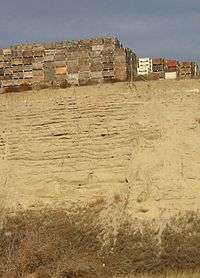  This photo shows a canyon cut into the surrounding flat soil with over 20 distinct horizontal layers of sediment, each clearly demarked from the layer below. Above the canyon lip a number of boxes used for transporting harvested apples are stacked; the size of these boxes indicates that the layers each are 0.5-1 meter in depth.