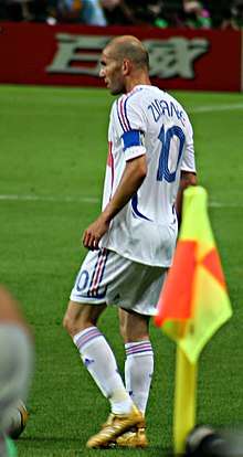 A balding man wearing a white shirt with white shorts and the number "10" and "Zidane" on the back.