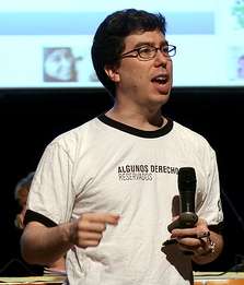waist high portrait wearing a T-shirt reading "Algunos Derechos Reservados", holding a microphone hand and a marker in the other