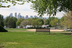 Photograph of a park bench and fields in the foreground and the City of Denver skyline in the background.