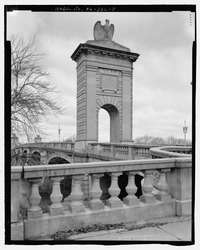 A stone arch with stone eagle is in the background near the entrance to a bridge that continues beyond it. A stone banister and railing in the foreground.