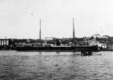 A black warship with two masts and a single funnel in port.