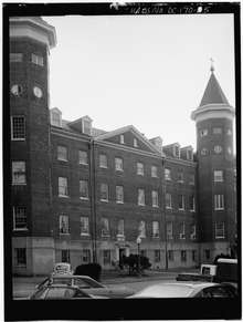 Black and white photograph of the façade brick building with two towers attached to the front. Roofs of cars are visible in the foreground