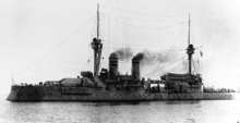 A large ship steaming, black smoke belching from her funnels, with many crewmen on the decks