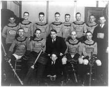 A black and white photo of ice hockey players sitting in two rows on a bench. Their jerseys feature the American flag on the front. Their coach is seated front row, center, with a young boy sitting in front of him.