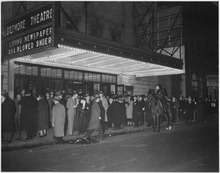 Black-and-white photo of a crowd on the sidewalk in front of a building with a lighted marquee saying 'Biltmore Theatre'.